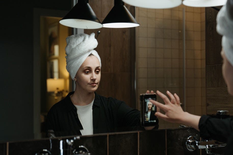 Girl with towel over hair in bathroom live streaming a video to her phone on the mirror